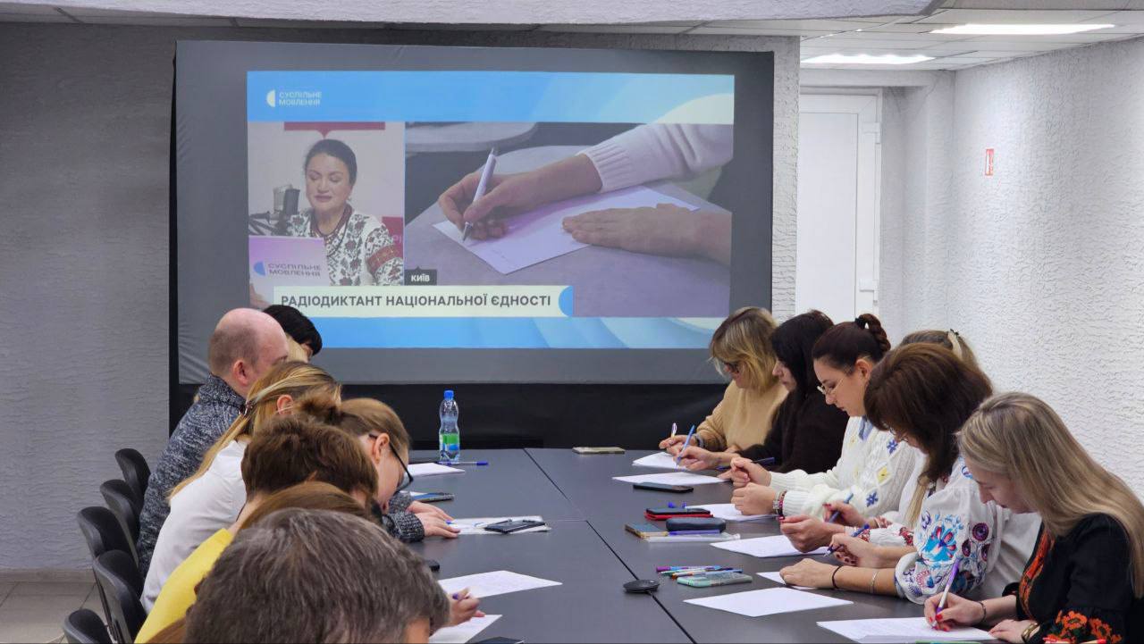 Група людей сидить за довгим столом і пише, беручи участь у "Радіодиктанті національної єдності", що транслюється на великому екрані на задньому плані. На екрані відображається відео з диктором (жінкою) та кадр із процесом писання. Деякі учасниці одягнені у вишиванки. На столі лежать папери, ручки та пляшка води.
