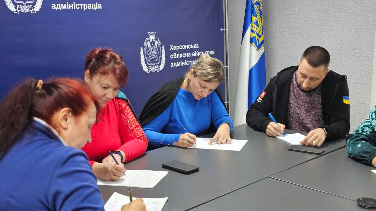 Четверо людей (троє жінок і один чоловік) сидять за столом і пишуть, зосередившись на паперах. На задньому плані розташований синій банер із логотипом і написом "Херсонська обласна військова адміністрація". Праворуч стоїть прапор із синьо-жовтими елементами.
