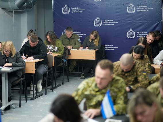 Група людей сидить за партами і пише у приміщенні для навчання. Серед учасників є люди у цивільному одязі та військовослужбовці у камуфляжній формі ЗСУ. На задньому плані розташований великий синій банер (пресвол) із логотипами та написом "Херсонська обласна військова адміністрація". На передньому плані видно розмиту фігуру людини у камуфляжі та мініатюрний синьо-білий прапорець.
