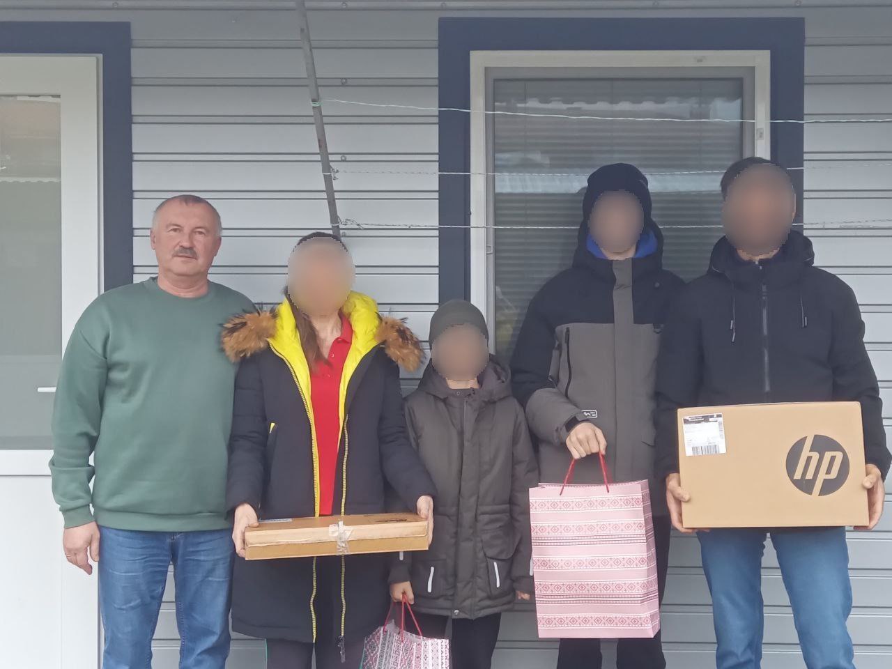 Груповий портрет п'яти осіб, які стоять перед фасадом будівлі, обшитим сірим сайдингом, і, ймовірно, отримують допомогу.  Зліва стоїть чоловік (можливо, волонтер або представник), одягнений у зелений светр і сині джинси, з вусами.  Праворуч від нього стоїть жінка, обличчя якої приховано. Вона одягнена у чорну зимову куртку з хутряним капюшоном та жовту кофту. Вона тримає в руках довгу, плоску картонну коробку.  У центрі стоїть дитина (хлопчик), також із прихованим обличчям, одягнена в теплу темну куртку, тримає в руках невеликий подарунковий пакет.  Праворуч стоять двоє старших юнаків чи чоловіків у темному зимовому одязі та шапках, обличчя яких приховані. Один із них тримає рожевий подарунковий пакет, інший — велику картонну коробку від ноутбука HP.  Уся група стоїть перед сірою стіною з білим віконним і дверним обрамленням. Сцена відображає передачу допомоги або подарунків.