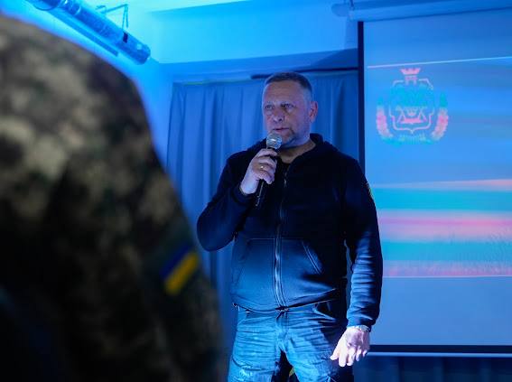 Чоловік середнього віку у темній кофті з капюшоном та джинсах стоїть, тримаючи мікрофон біля рота, перед екраном із логотипом на синьому тлі.