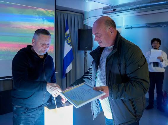 Двоє чоловіків стоять у приміщенні з синім освітленням. Чоловік ліворуч у темній кофті дістає щось зі світлого паперового пакета, який стоїть на столі або підставці, тоді як чоловік праворуч у темній куртці тримає в руках рамку з грамотою або сертифікатом. На задньому плані видно прапор (синьо-білий) на підставці, динамік та ще одного чоловіка, який тримає стопку паперів.