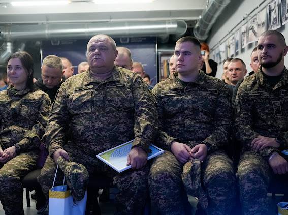 Група військовослужбовців Збройних Сил України, включно з однією жінкою, сидять у приміщенні та слухають виступ. Усі одягнені в камуфляжну форму. Чоловік у центрі на передньому плані, із помітною статурою, тримає рамку з грамотою або сертифікатом. На задньому плані видно стіну, прикрашену фотографіями або документами.