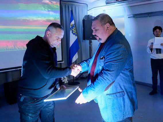 Двоє чоловіків потискають один одному руки у приміщенні з синьо-блакитним освітленням. Чоловік ліворуч у темній кофті тримає рамку з грамотою чи сертифікатом. Чоловік праворуч, більшої статури, одягнений у синій піджак і сорочку з червоними елементами. На задньому плані видно екран із кольоровим зображенням, прапор (синьо-білий), динамік та ще одного чоловіка, який тримає папери.