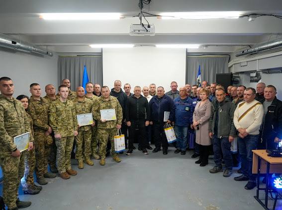 Груповий знімок близько тридцяти чоловіків та двох жінок, які стоять у приміщенні. Більшість людей ліворуч одягнені у військову камуфляжну форму (ЗСУ), деякі тримають рамки з грамотами та паперові пакети. Люди праворуч одягнені у цивільний одяг. На задньому плані видно великий білий екран, два прапори (синьо-жовтий і синьо-білий), динамік та проектор на стелі.