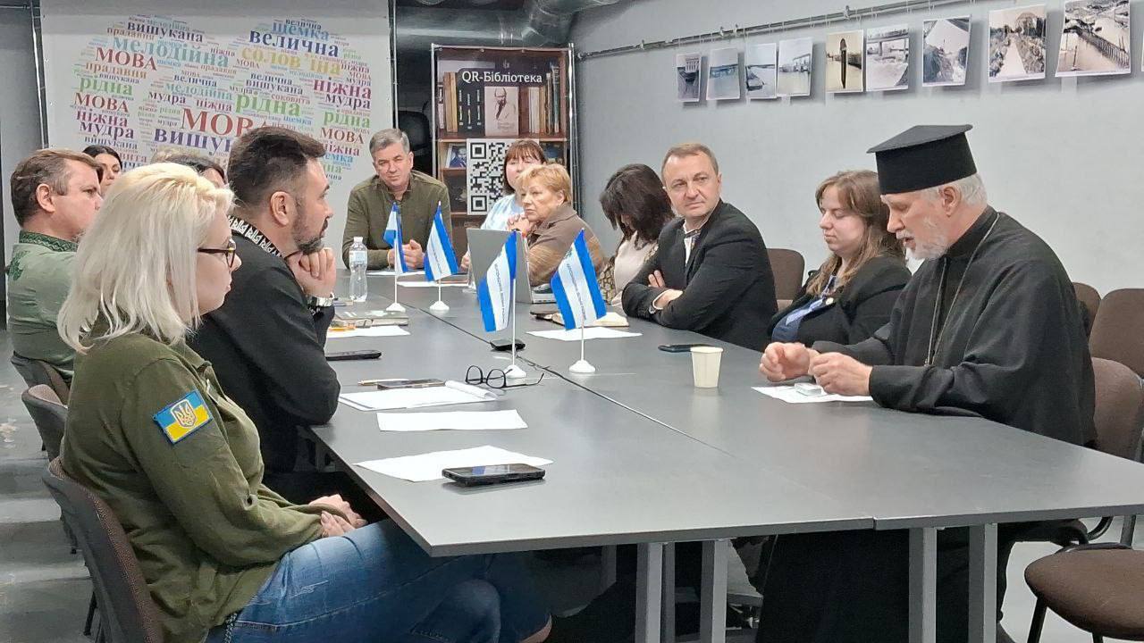 Група з приблизно десяти осіб сидить за великим прямокутним столом під час зустрічі. Серед учасників — жінка на передньому плані з білим волоссям, одягнена у військову куртку з прапором України на рукаві; чоловік у рясі та скуфії (праворуч); та інші люди у цивільному або військовому одязі. На столі стоять мініатюрні прапорці (синьо-білі) та розкладені окуляри, телефони та кава. На стінах видно плакат із написом «QR-Бібліотека» та слова, що стосуються української мови ("мова", "рідна", "велична"), а також низку чорно-білих фотографій.