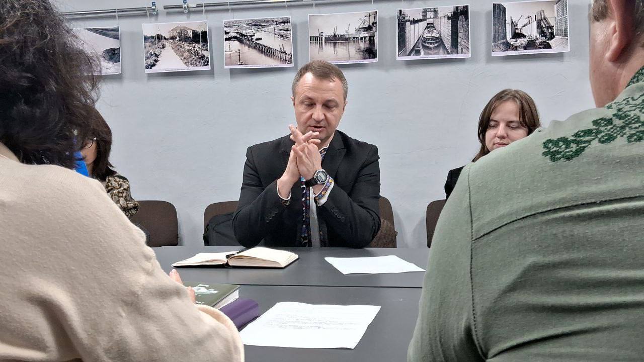 Чоловік у чорному костюмі та краватці сидить за столом, склавши руки в замок, і, ймовірно, говорить або уважно слухає. На столі перед ним лежить відкритий блокнот і аркуші паперу. Двоє інших учасників зустрічі, включно з жінкою праворуч, сидять поряд. На передньому плані видно частину голови та плеча іншої особи. На стіні за чоловіком висять п'ять чорно-білих фотографій із зображеннями, схожими на портові або міські пейзажі.