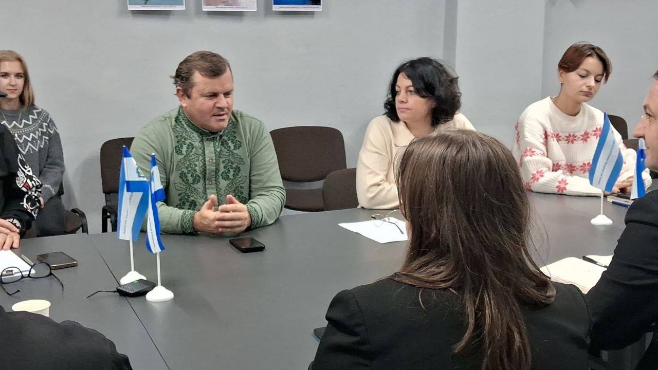 Група людей сидить за столом під час ділової зустрічі. У центрі композиції чоловік у світло-зеленій вишиванці, який активно говорить, склавши руки. Поруч із ним сидять дві жінки (одна у світлому светрі, інша — у білому/бежевому). На столі стоять два мініатюрні прапорці (синьо-білі) і лежать мобільні телефони та окуляри. 