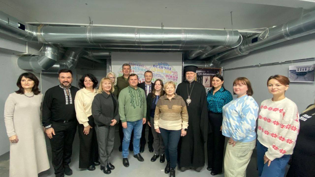 Груповий знімок близько п'ятнадцяти осіб, які стоять у приміщенні. Серед них — кілька чоловіків у вишиванках, чоловік у чорній рясі та скуфії, а також жінки у різному цивільному одязі. На задньому плані розташований великий екран із написами, пов'язаними з українською мовою (зокрема, слова "велична", "ніжна", "мудра" тощо), а також плакат із написом "QR-Бібліотека". 