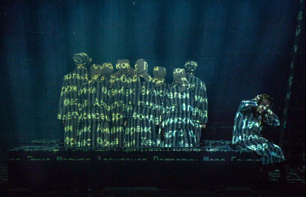 Зображення є кадром з театральної вистави, що демонструє групу акторів на темній сцені.  На сцені вісім осіб стоять у центрі, обернувшись спиною до глядача. Вони одягнені у смугасті роби, що нагадують тюремну форму або форму в'язнів концтабору, а голови покриті хустками або шапками.  На їхні роби та на стіну позаду проєктуються жовто-зелені літери та слова кирилицею, які роблять їх напівпрозорими та примарними. Серед чітко видимих слів можна розпізнати: 