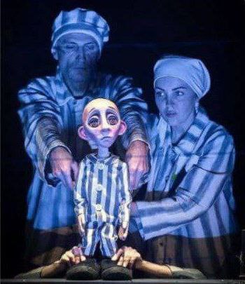 Зображення є кадром з театральної вистави, на якому зображено двох акторів, які маніпулюють маріонеткою.  Актори (чоловік і жінка) стоять на темному тлі і одягнені у смугасті роби та головні убори, схожі на тюремну форму або одяг в'язнів концтабору, освітлені синім світлом. Вони дивляться прямо вперед.  Лялька-маріонетка розташована по центру між ними. Це лялька хлопчика із сумним, великими очима та виразним обличчям. Вона також одягнена у смугастий одяг.  Руки акторів тримають ляльку за плечі та з боків. Унизу кадру видно ще дві руки в чорному одязі, які, ймовірно, також керують лялькою.  Зображення передає драматичну та зворушливу сцену, зосереджену на темі дитинства, вразливості та поневолення.