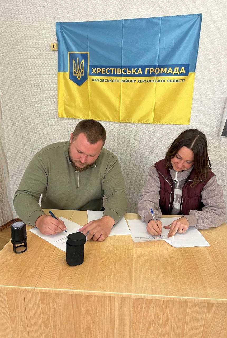 Зображення, зняте в приміщенні, показує чоловіка та жінку, які сидять за дерев'яним столом і підписують документи. Чоловік сидить ліворуч, одягнений у зелений светр або кофту, з бородою, і заповнює папери ручкою. Жінка сидить праворуч, одягнена у світлий светр та бордовий жилет, також підписує документи. На столі видно невелику чорну колонку та штамп. На стіні за ними висить прапор України (синьо-жовтий) з нанесеним на нього текстом: ХРЕСТІВСЬКА ГРОМАДА КАХОВСЬКОГО РАЙОНУ ХЕРСОНСЬКОЇ ОБЛАСТІ Зображення, ймовірно, фіксує момент оформлення важливих документів у представництві місцевої влади.