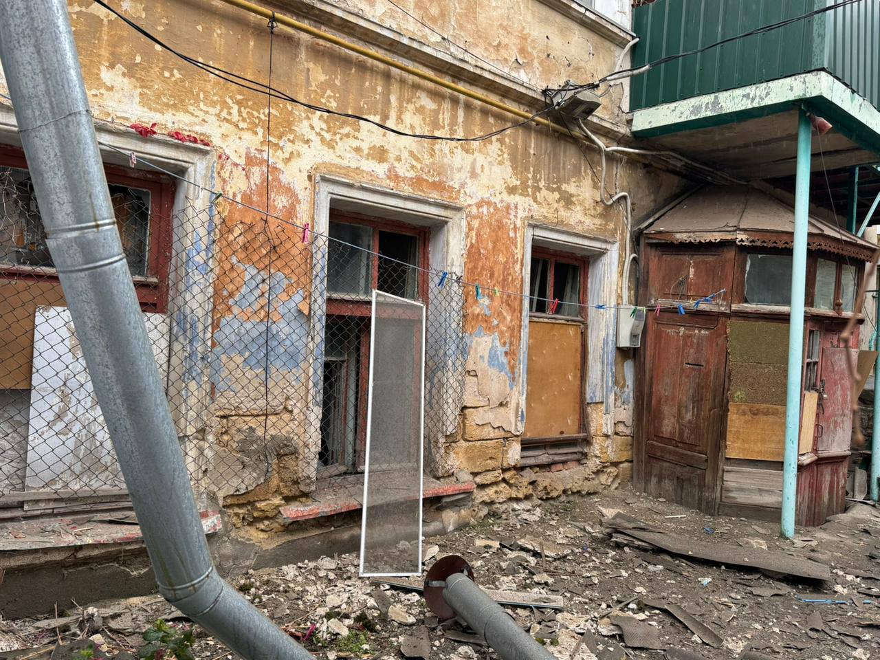 Зображення демонструє сильно пошкоджений, захаращений внутрішній дворик або задній фасад старої будівлі. Стіни мають значні руйнування: облущена фарба, оголені цегла та каміння, сліди обвітрювання та обстрілів.  На фасаді видно три віконні прорізи з розбитими шибками: один ліворуч частково закритий сіткою-рабицею, натягнутою перед вікнами, а два вікна праворуч забиті фанерою чи дошками. По центру перед вікнами на землі стоїть довгий прямокутний шматок скла або пластику.  Праворуч розташовані старі дерев'яні двері та невелика прибудова чи веранда з іржавим металевим дахом. Зверху видно невеликий зелений металевий балкон.  На передньому плані видно велику металеву трубу, що йде навскіс через кадр, а земля вкрита сміттям, уламками та ґрунтом. Загальний вигляд передає атмосферу занедбаності та руйнування внаслідок військових дій.