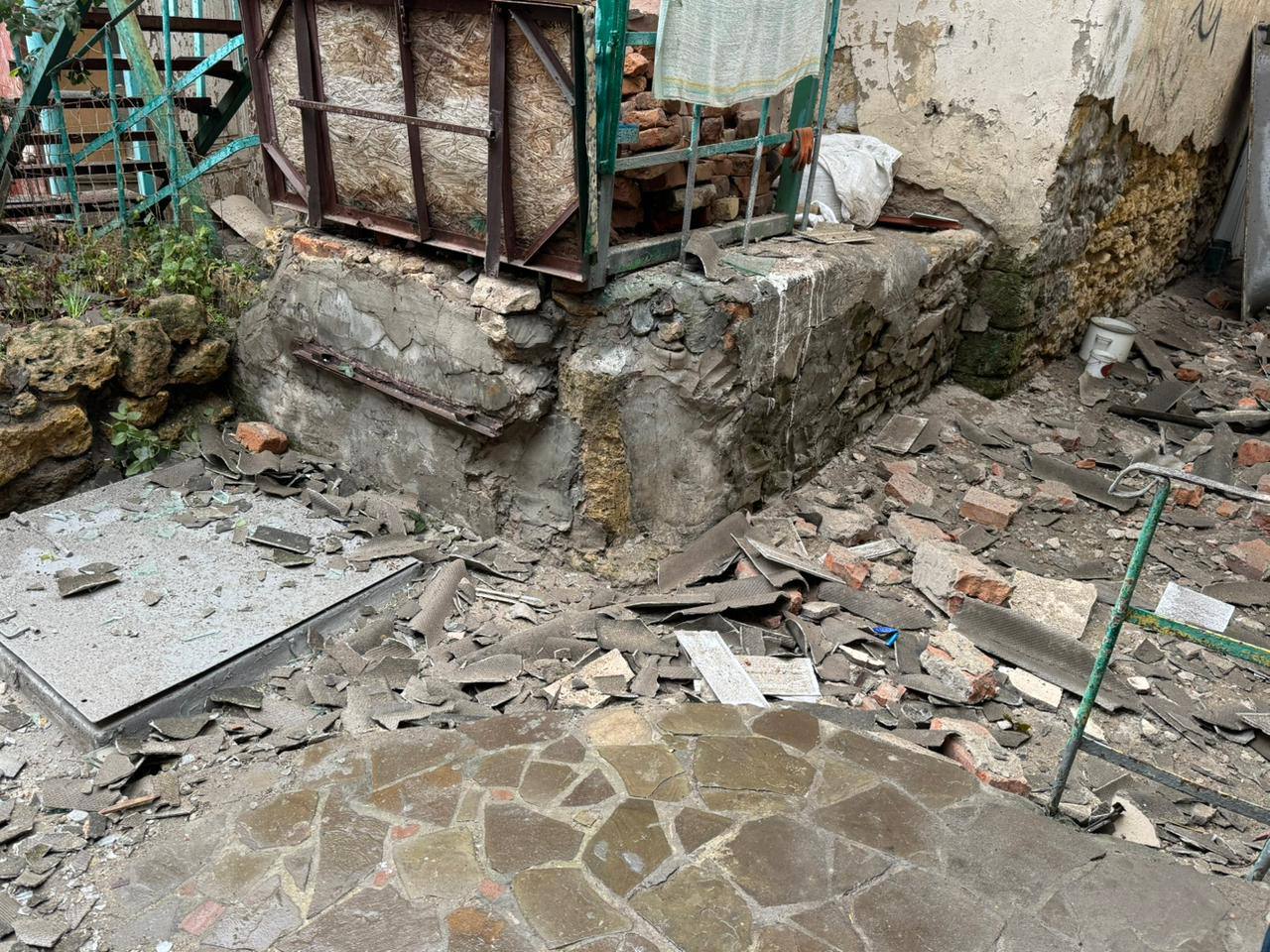 Зображення демонструє руйнування у внутрішньому дворику чи біля фундаменту будівлі. На передньому плані видно частину вимощеної кам'яної підлоги (мозаїка з нерівних кам'яних плиток).  Решта простору вкрита великою кількістю будівельного сміття, уламків шиферу, цегли та бетону, що свідчить про значні пошкодження.  У центрі та на задньому плані розташований масивний, напівзруйнований фундамент або цоколь будівлі з обшарпаним бетоном і цеглою. Зверху на фундаменті стоїть імпровізована конструкція, схожа на закритий металевий ящик або господарську прибудову з іржавими металевими каркасами та частинами, зашитими фанерою/дошками. Стіна будівлі позаду має сліди сильного пошкодження. Ліворуч видно зелену рослинність, а праворуч на смітті лежить зелена металева драбина або каркас. Загальний вигляд передає наслідки обстрілу та повне руйнування елементів споруди.
