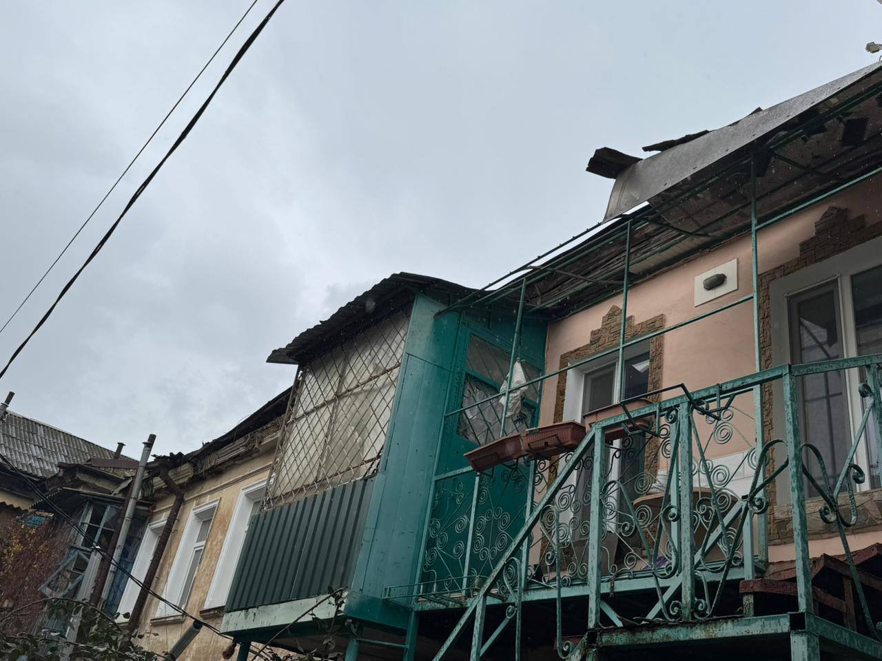 Зображення, зняте з нижнього ракурсу на тлі сірого неба, демонструє пошкоджені балкони та дахи житлового будинку старої забудови.  На передньому плані видно металевий балкон із різьбленою балюстрадою, пофарбованою у зелений (бірюзовий) колір. З балкона ведуть зовнішні металеві сходи.  Праворуч розташована стіна будівлі рожевого або блідо-персикового кольору, з вікном. Ліворуч — частина стіни жовтого відтінку. Між ними виділяється засклений балкон або прибудова насиченого бірюзово-зеленого кольору, зі значними пошкодженнями даху та елементів обшивки, зокрема, видно вибите скло або пластик із внутрішньою сіткою.  Дахи над усіма частинами будівлі мають ознаки руйнування, відсутні фрагменти покрівлі. Уздовж верхньої частини кадру тягнуться дроти електропередач. Зображення передає атмосферу занедбаності та шкоди, заподіяної будівлі.