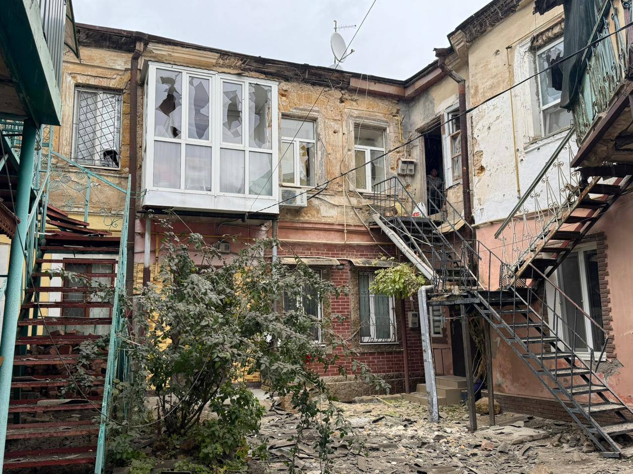 Зображення демонструє пошкоджений внутрішній дворик старого житлового будинку з численними ознаками руйнування.  У центрі кадру знаходиться засклений балкон на другому поверсі з білою рамою, скло якого повністю розбите, утворюючи великі тріщини та отвори. Поруч із балконом видно інші вікна, одне з яких має решітку.  Фасади будівлі знаходяться у поганому стані: стіни мають облущену фарбу, тріщини та сліди обстрілів. Нижній поверх частково облицьований червоною цеглою.  У дворику розташовані дві старі, іржаві металеві зовнішні сходи, що ведуть на другий поверх. Одна з них, ліворуч, має сходинки зі значним нахилом.  На землі у дворику багато сміття, уламків та будівельних матеріалів. У центрі росте великий кущ. Зверху на даху/стіні видно невелику супутникову антену. Загальний вигляд передає значні руйнування будинку внаслідок військових дій.