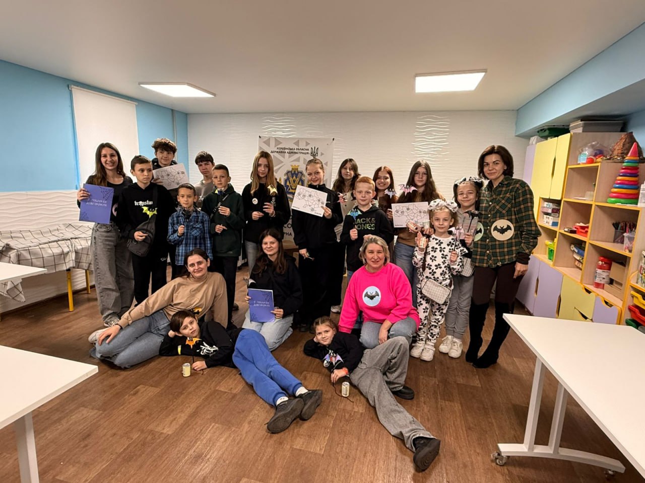 Зображення демонструє групове фото дітей та дорослих у світлій кімнаті, ймовірно, в дитячому центрі або класі. Стіни кімнати частково пофарбовані у блакитний колір.  У групі приблизно 15 дітей різного віку та п'ять дорослих (переважно жінок). Усі учасники позують, усміхаються, деякі тримають у руках сертифікати, малюнки або невеликі подарунки (зірки).  Деякі особи стоять у задньому ряду, а інші сидять або лежать на дерев'яній підлозі у передньому ряду. Дорослі одягнені у повсякденний одяг, одна жінка праворуч одягнена в картату сорочку та чоботи, інша в центрі – у яскраво-рожевий светр.  На задньому плані, на стіні, висить плакат із символікою та текстом, який частково видно: 