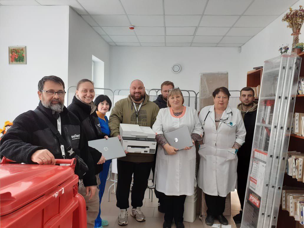 Група з восьми людей (п'ять чоловіків і три жінки) стоїть у світлому приміщенні, схожому на медичний заклад або офіс.  На передньому плані зліва чоловік у чорній куртці та окулярах, з бородою, спирається на великий червоний контейнер або візок.  Поруч із ним усміхнена жінка у чорній куртці.  В центрі стоїть бородатий чоловік у куртці кольору хакі, який тримає чорно-білий принтер, а також тримає ноутбук.  Праворуч від нього чоловік стоїть трохи позаду.  Дві жінки у білих халатах (можливо, лікарки чи медсестри) стоять з правого боку. Жінка ближче до центру тримає ноутбук, а інша жінка у халаті з фонендоскопом на шиї.  Скраю праворуч стоїть ще один чоловік.  Загалом, люди тримають принаймні три ноутбуки та один принтер, що може свідчити про передачу обладнання.  Праворуч видно високі металеві стелажі з книгами чи документами та складену драбину.  На стіні зліва висить невелика картина з квітами.