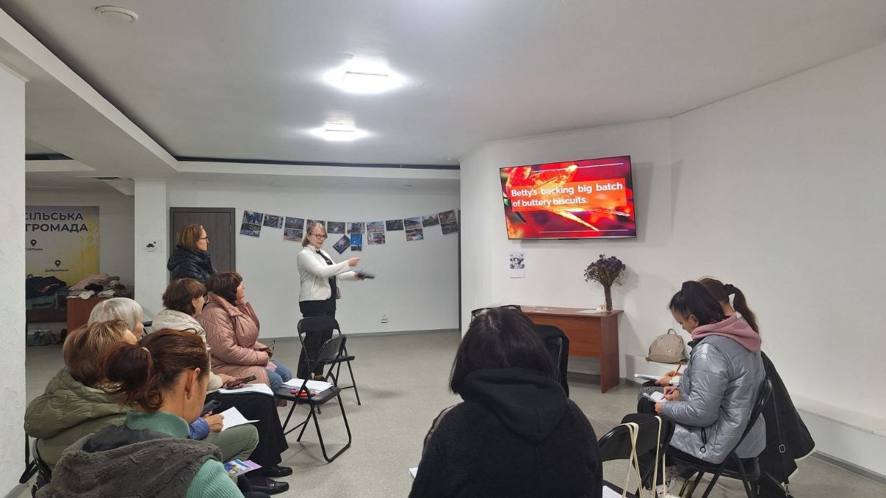 Фотографія закритого приміщення, ймовірно, класу чи лекційної кімнати, де відбувається презентація. Жінка в світлому піджаку стоїть посередині, обличчям до аудиторії, і жестикулює, пояснюючи щось. Приблизно десяток жінок різного віку сидять на стільцях, уважно слухають і роблять нотатки. На стіні праворуч висить телевізійний екран, на якому відображається текст англійською: 