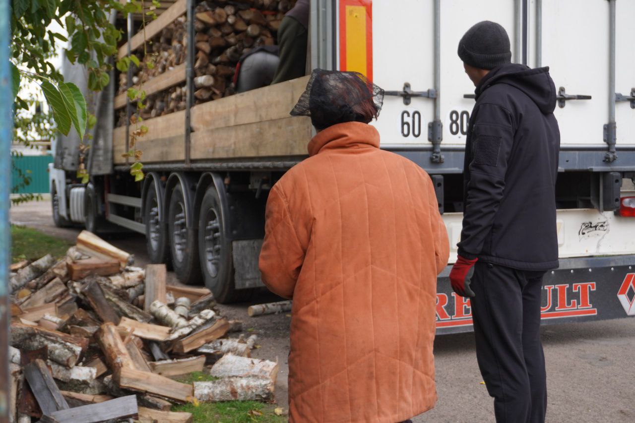 Зображення демонструє момент розвантаження дров із великої вантажівки.  На задньому плані видно частину довгої вантажівки-причепа з високими дерев'яними бортами, заповненої дровами. На білій задній частині причепа помітні написи 