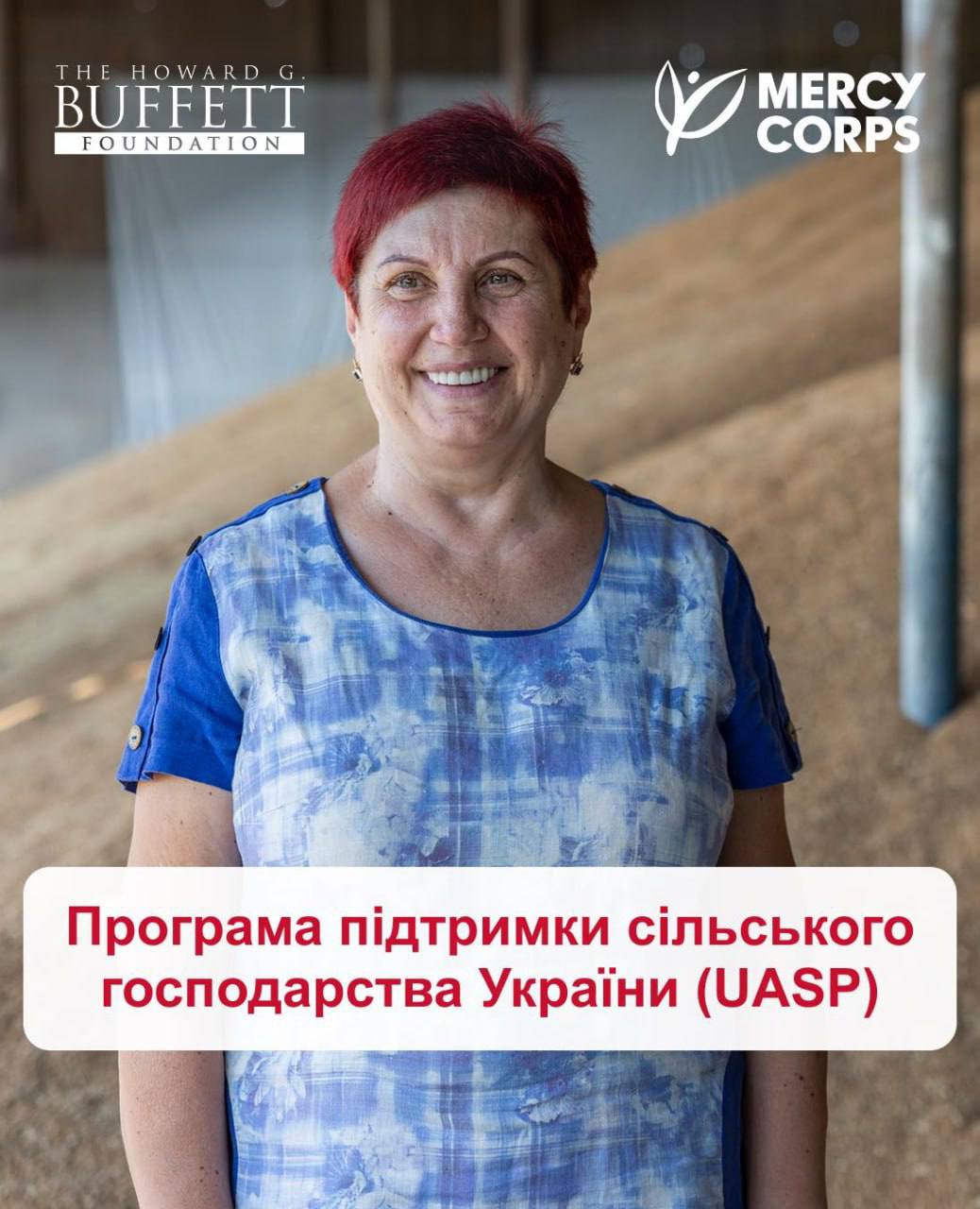 Вертикальне рекламне зображення, присвячене аграрній допомозі. У центрі кадру крупним планом зображено усміхнену жінку середнього віку з коротким рудим волоссям, одягнену в синьо-білу картату блузку. Жінка стоїть на тлі зерносховища або елеватора, де видно велику кількість насипаного зерна. У верхніх кутах розташовані логотипи: Ліворуч: THE HOWARD G. BUFFETT FOUNDATION (Фонд Говарда Г. Баффета). Праворуч: MERCY CORPS (Мерсі Корпс). У нижній частині зображення накладено червоний прямокутник із великим білим текстом: Програма підтримки сільського господарства України (UASP) Зображення анонсує спільну програму підтримки агросектору від згаданих організацій.