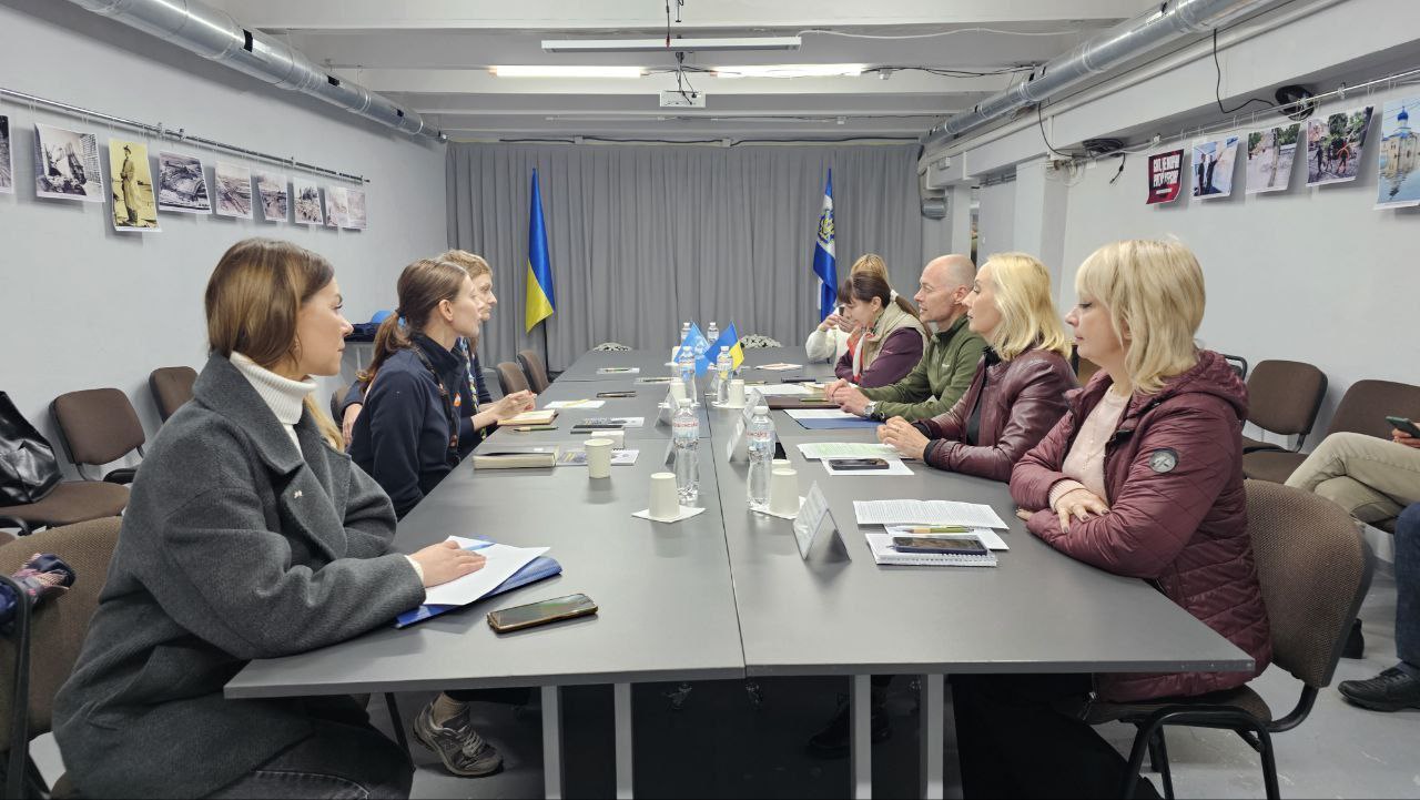 Робоча зустріч за довгим столом, в якій беруть участь вісім осіб. За столом сидять керівники профільних структур Херсонської ОВА, представники Херсонської МВА та міських служб, які проводять переговори з представниками Фонду ООН у галузі народонаселення (UNFPA). На задньому плані видно державний прапор України та прапор міжнародної організації. На столі розташовані пляшки з водою та робочі документи.