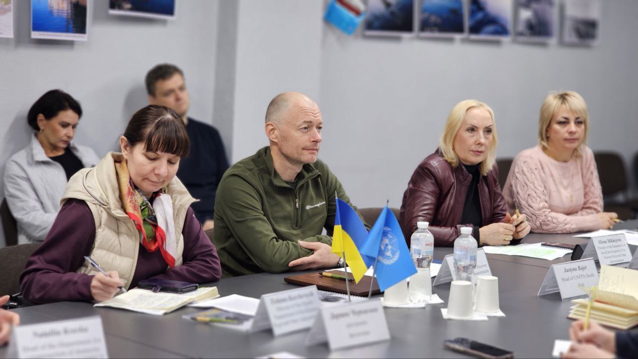 Фотографія крупним планом учасників робочої зустрічі керівників профільних структур Херсонської ОВА, Херсонської МВА та міських служб з представниками Фонду ООН у галузі народонаселення (UNFPA). На передньому плані видно чотирьох учасників, які сидять за столом: жінка в жилетці робить нотатки, чоловік у формі кольору хакі уважно слухає, та дві жінки дивляться на спікера. Перед ними стоять маленькі прапорці України та ООН (блакитний прапор із символікою).