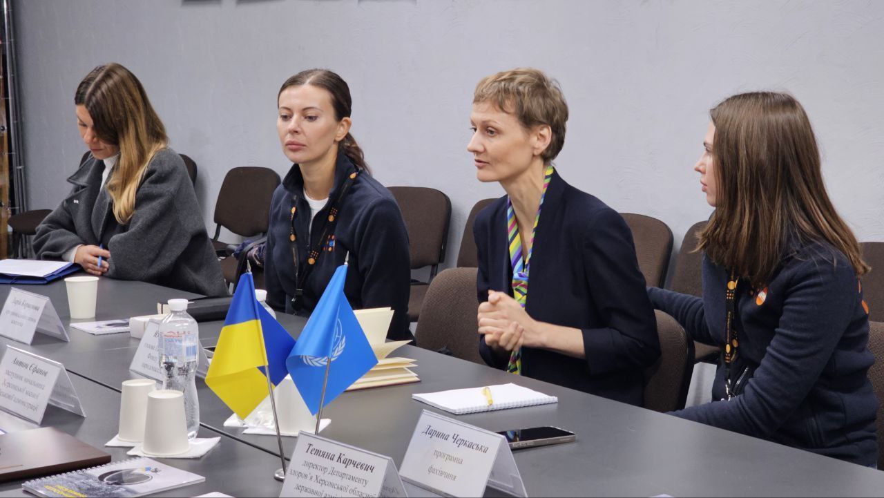 Крупний план чотирьох учасниць робочої зустрічі керівників профільних структур Херсонської ОВА/МВА та представників міських служб з представниками Фонду ООН у галузі народонаселення (UNFPA). Жінки сидять за столом; та, що знаходиться у центрі (в темному піджаку з різнобарвною хусткою/краваткою), активно говорить і жестикулює. На столі перед ними стоять маленькі прапори України та ООН, а також іменні таблички.