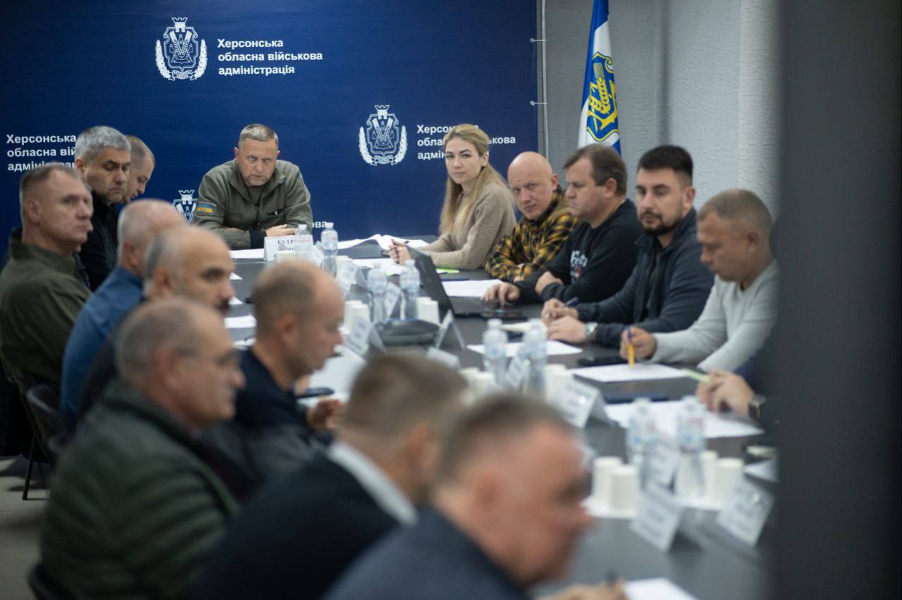 Короткий альтернативний текст:  Нарада в приміщенні Херсонської обласної військової адміністрації. Група з понад десяти людей сидить за столом на тлі синьої стіни з гербом ОВА. У центрі сидить чоловік у військовій формі, який керує зустріччю.