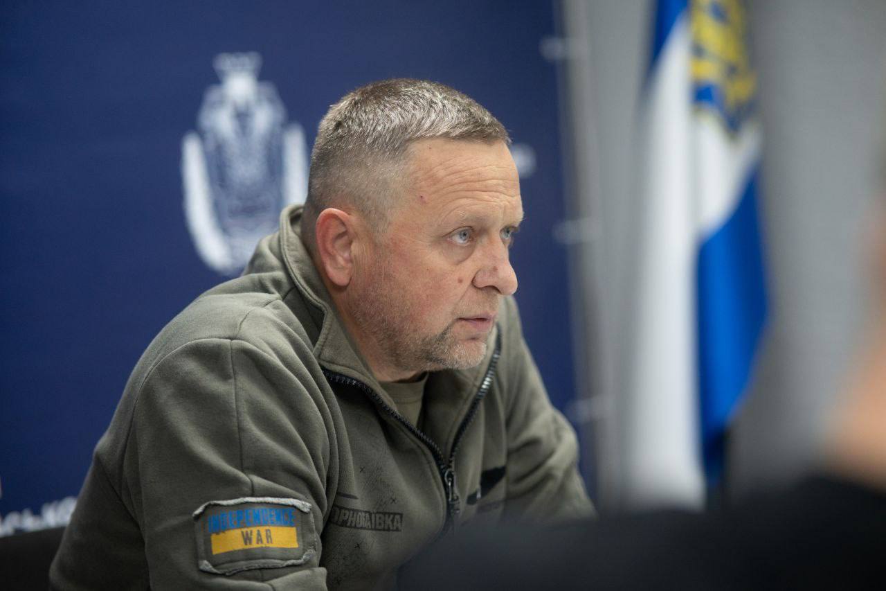 Крупний план чоловіка середнього віку з короткою стрижкою, який сидить на нараді. У нього світлі очі та зосереджений вираз обличчя. Чоловік одягнений у військову флісову кофту кольору хакі (оливковий).