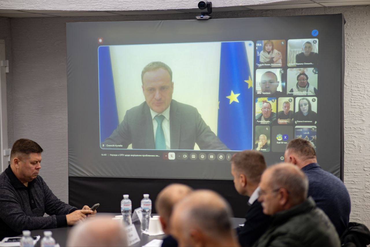 Нарада, де учасники в кімнаті (на передньому плані) дивляться на великий екран. На екрані відбувається відеоконференція, головний спікер – Олексій Кулеба, який виступає на тлі українського прапора та прапора ЄС.