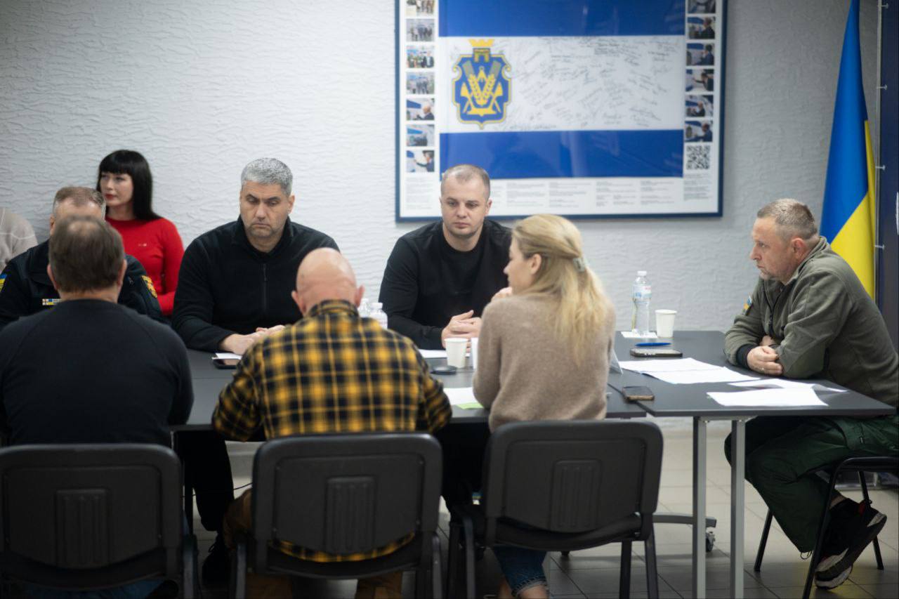 Фрагмент наради в Херсонській ОВА: учасники сидять за столом на тлі плаката з гербом Херсонщини та українським прапором. На фото видно чоловіка у військовій формі (праворуч) та двох жінок, одна з яких спілкується з чоловіком навпроти.