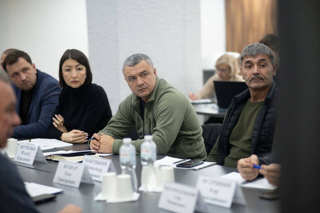 Група учасників наради, що сидять за столом. Серед них: чоловік у зеленій флісовій кофті в центрі, жінка в чорному светрі ліворуч, та чоловік із вусами та сивиною праворуч.