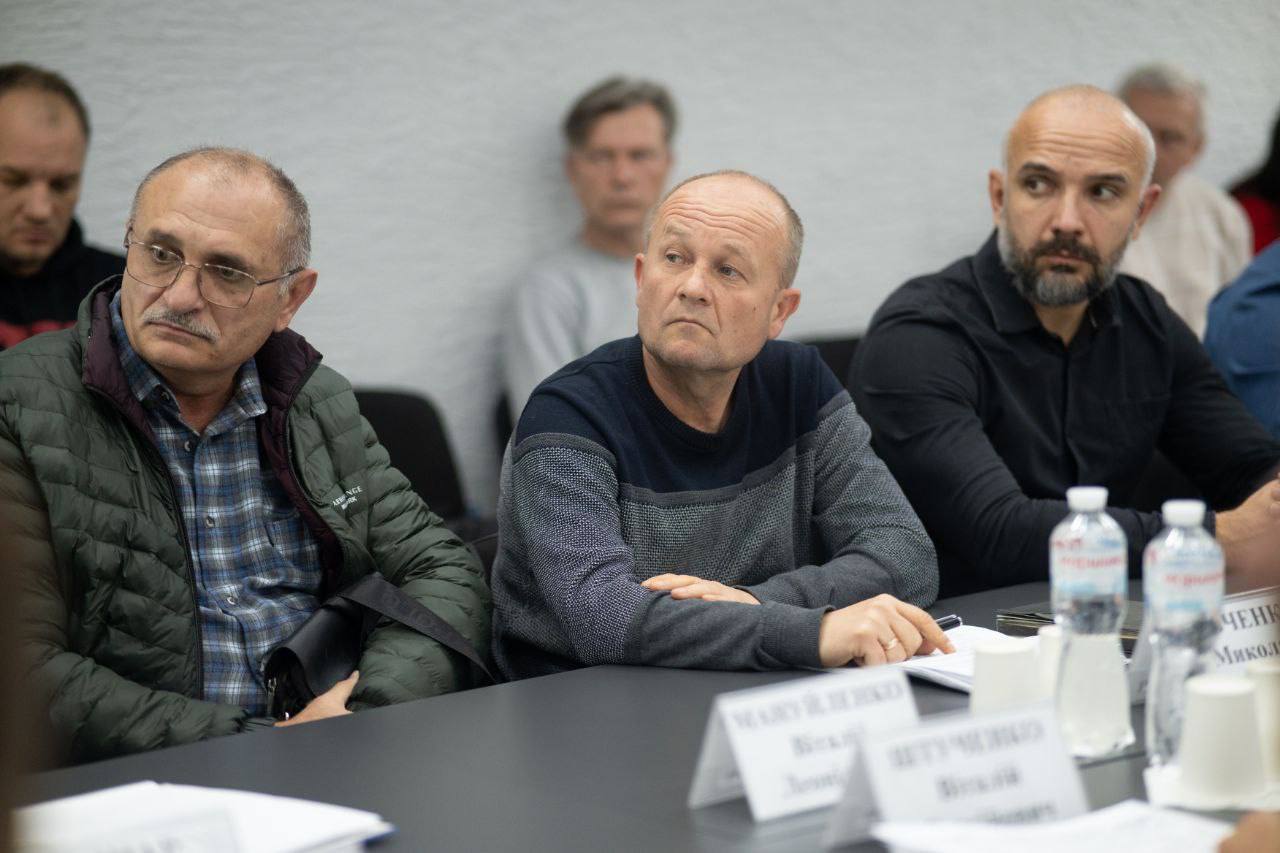 Крупний план трьох чоловіків за столом на нараді, зокрема, лисого чоловіка в окулярах у зеленій куртці (ліворуч) та чоловіка з бородою (праворуч).