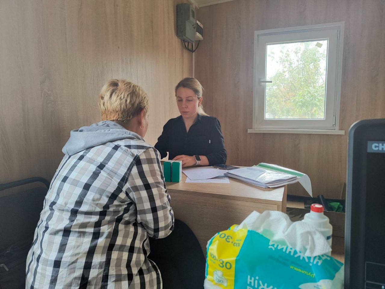 Жінка-консультантка (або медичний працівник) в темній сорочці сидить за столом і проводить консультацію жінці в картатій сорочці. Дія відбувається у приміщенні з дерев'яною обшивкою стін та невеликим вікном. На столі лежать документи, а на передньому плані видно пакет з гігієнічними засобами.