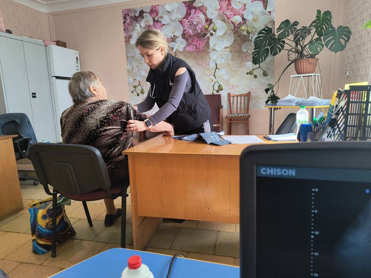 Медичний працівник (жінка у чорній жилетці зі стетоскопом) вимірює артеріальний тиск літній жінці-пацієнтці у кабінеті. На задньому плані видно квіткове панно, вазонну рослину та медичне/офісне приладдя. У нижньому правому куті частково видно екран ноутбука з написом 