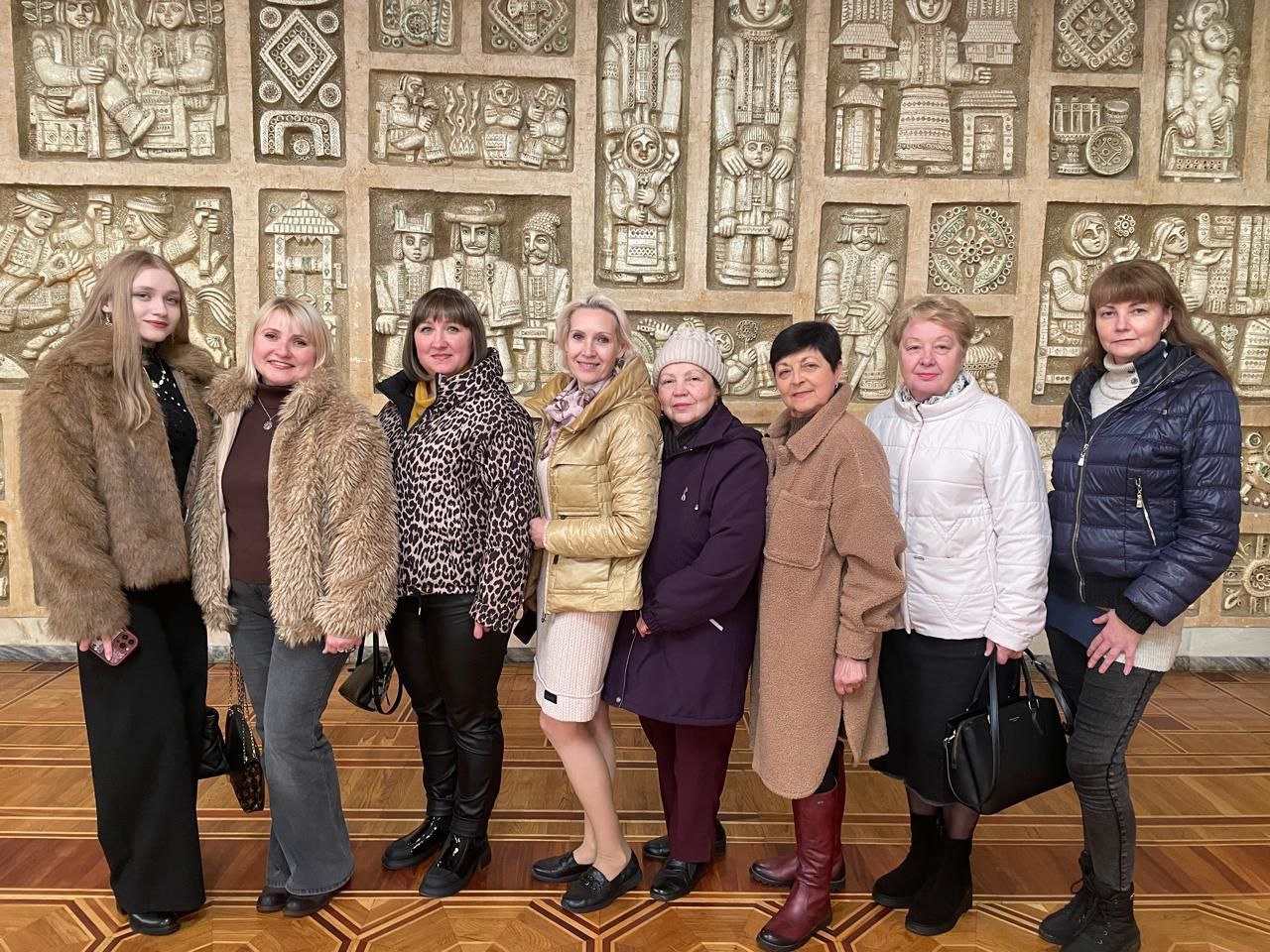 Груповий портрет восьми жінок, які стоять поруч і посміхаються перед великою, детально різьбленою або ліпною стіною.  На передньому плані: Жінки різного віку одягнені в зимовий або осінній одяг, включаючи хутряні пальта бежевих відтінків, світлий пуховик, темно-синю куртку, а також довгі пальта коричневих та фіолетових кольорів. Вони стоять у приміщенні з паркетною підлогою з геометричним візерунком.  На задньому плані: Стіна повністю покрита рельєфним панно світлого кольору (можливо, гіпс або камінь), що зображує стилізовані фігури людей в традиційному або історичному одязі та орнаменти, ймовірно, в українському народному або модерністському стилі. Це надає фону глибокої текстури та культурного значення.