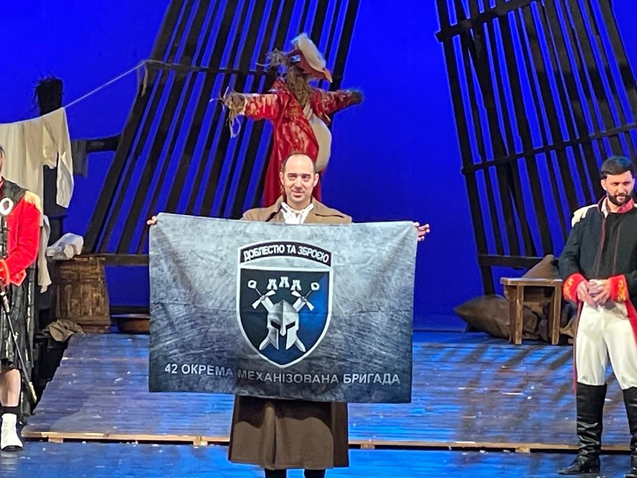 На сцені театру або іншого культурного закладу чоловік у коричневому вбранні (схожому на історичний костюм або частину декорації) тримає великий прапор 42 окремої механізованої бригади. На прапорі зображений герб бригади – щит із двома схрещеними мечами та лицарським шоломом, а також напис 