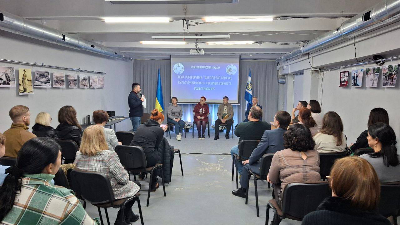 Публічний захід або лекція у приміщенні. Спікер стоїть біля групи людей, що сидять на сцені, перед аудиторією. На екрані видно тему презентації, а по боках сцени розташовані прапори України.