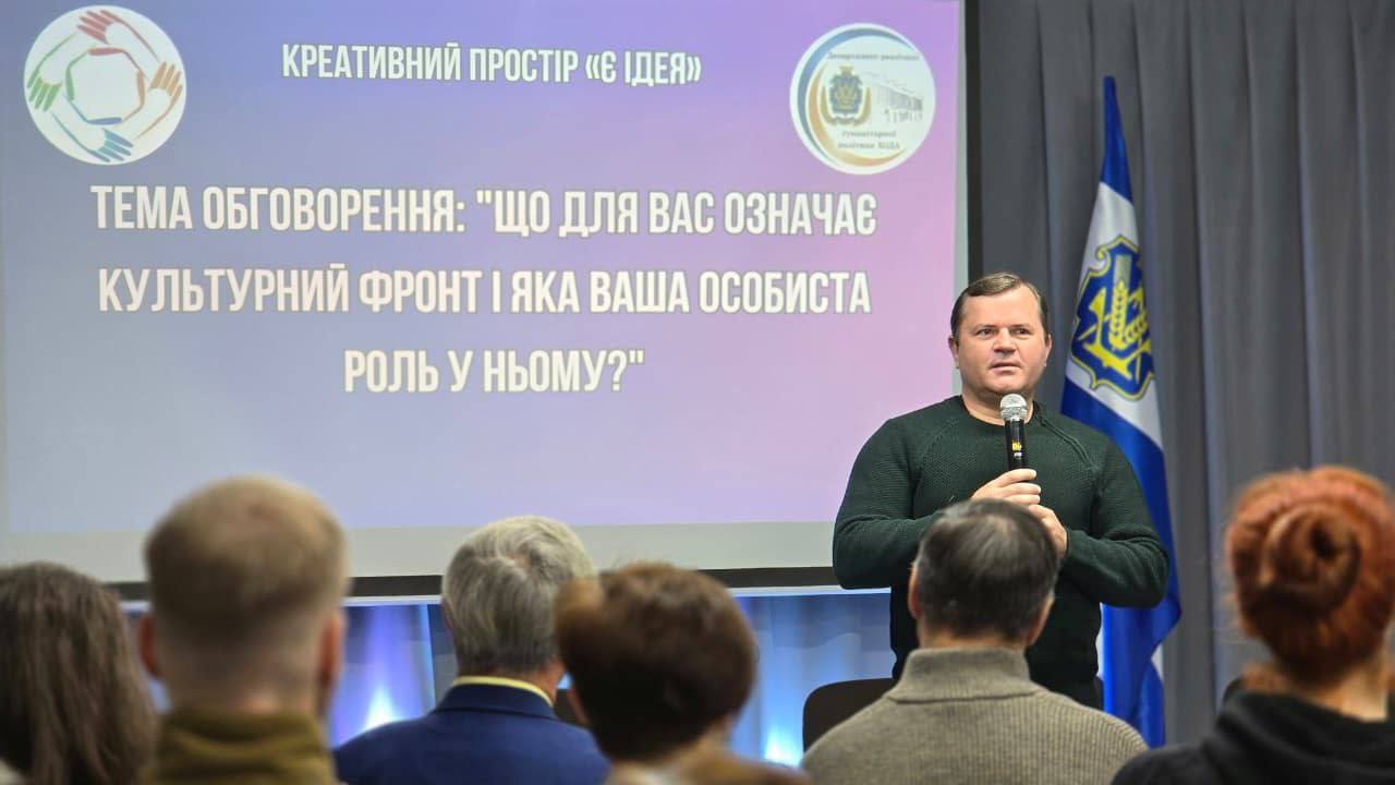 Чоловік у темно-зеленому светрі виступає перед аудиторією, тримаючи мікрофон. За ним — екран із текстом: 
