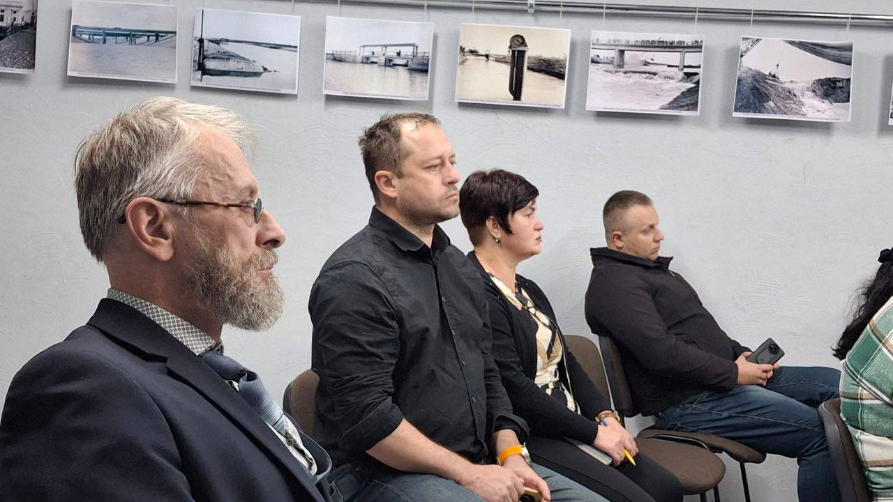 Четверо людей сидять у ряду, слухаючи виступ, на тлі стіни з чорно-білими фотографіями. На передньому плані — чоловік у окулярах із сивою бородою та чоловік у чорній сорочці.