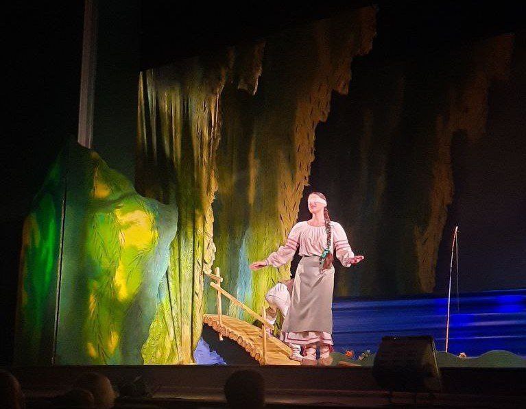 Акторка у світлому сценічному костюмі з пов'язкою на очах стоїть на сцені, простягаючи руки, на тлі декорацій, що імітують печеру та воду з містком.
