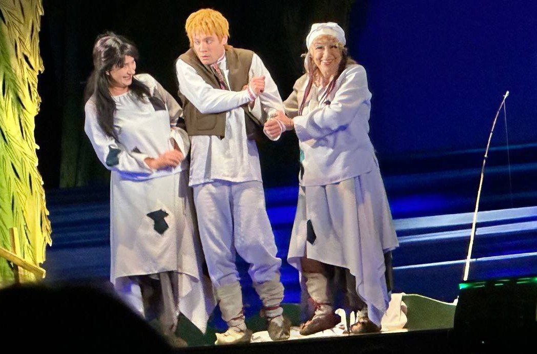 Троє акторів у традиційному сценічному одязі стоять на театральній сцені. По центру — чоловік із рудим волоссям, якого тримають за руки дві жінки по обидва боки.