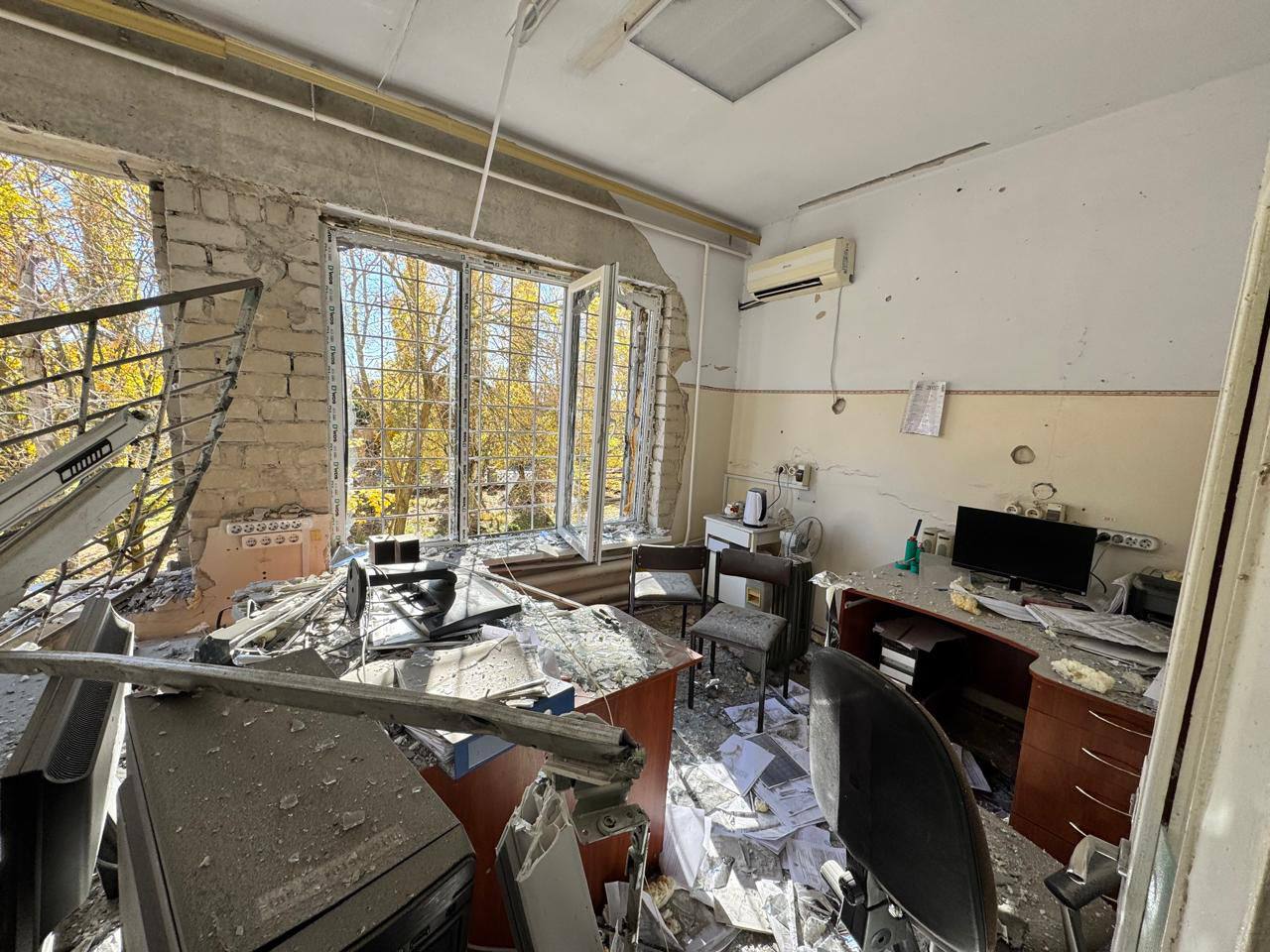 Зруйноване офісне приміщення після обстрілу. Стіни пошкоджені, вікна вибиті, меблі (столи, стільці) та офісна техніка завалені уламками та сміттям.