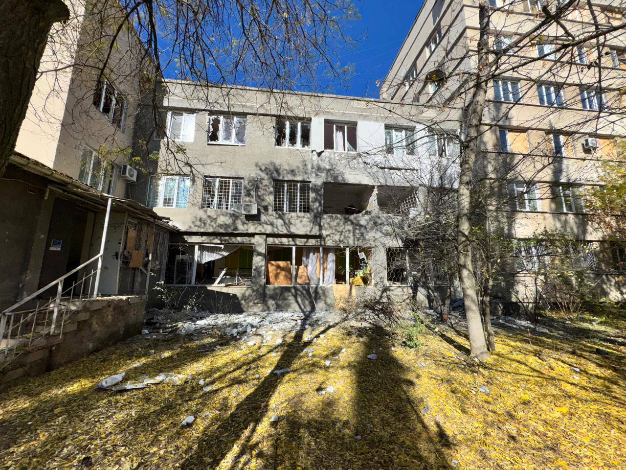 Зовнішній вигляд багатоповерхового житлового будинку з видимими пошкодженнями від обстрілу: вибиті вікна та зруйновані балкони. Земля перед будинком вкрита жовтим опалим листям та уламками.