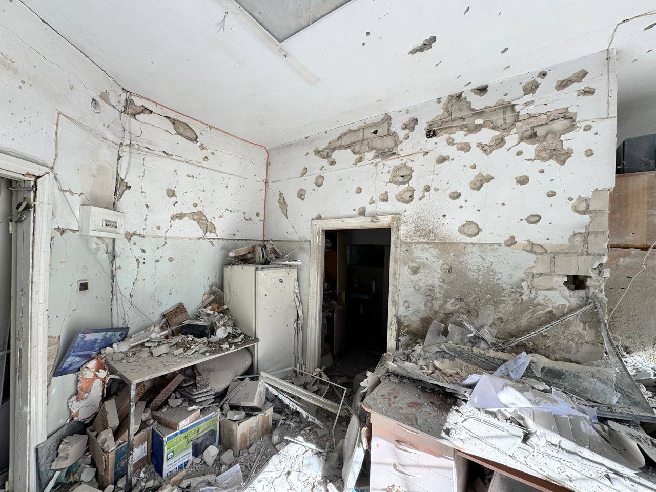 Сильно пошкоджена кімната або офіс. Стіни вкриті численними отворами та тріщинами від осколків, у приміщенні багато будівельного сміття, уламків меблів та техніки.
