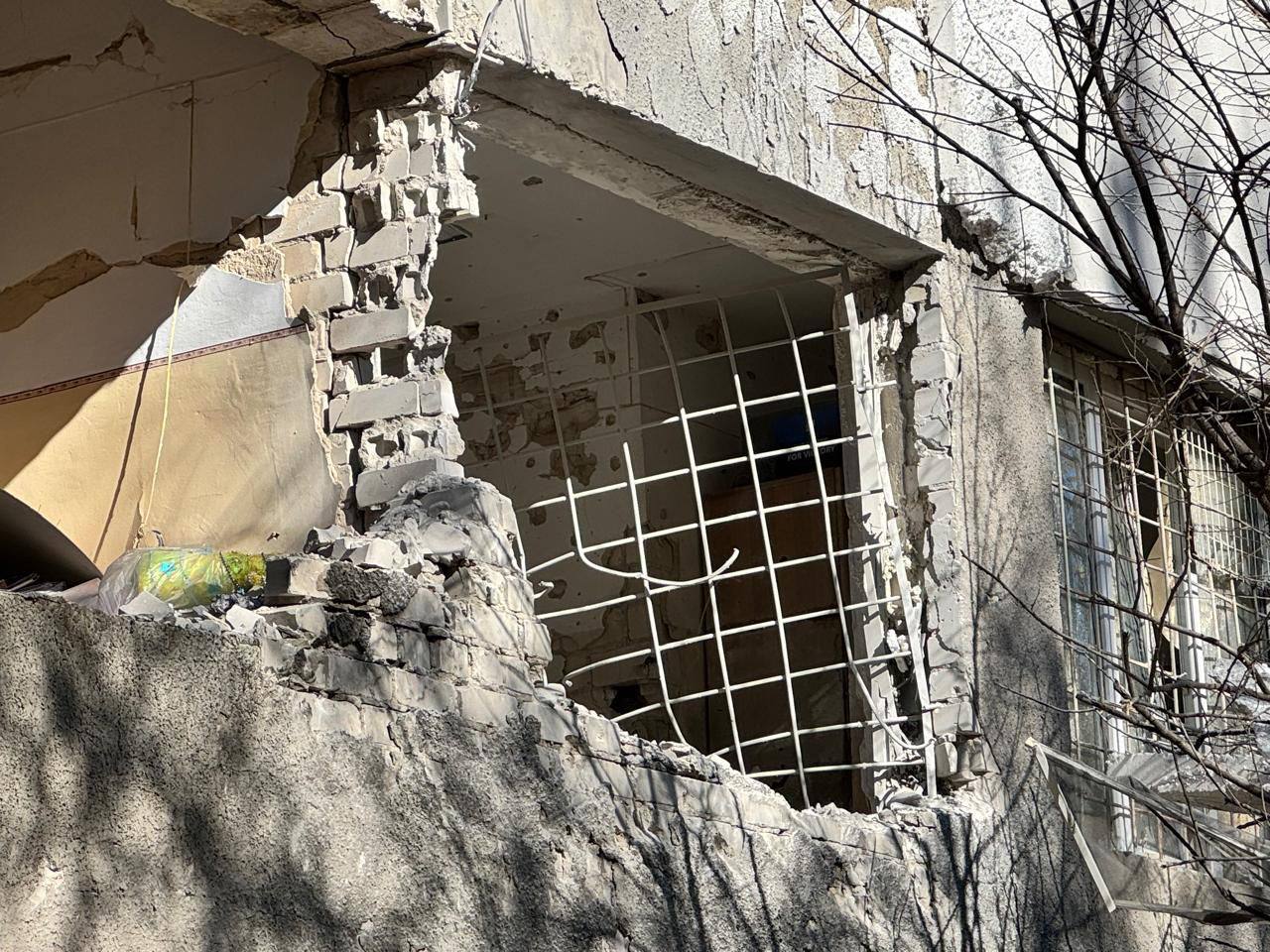 Зруйнована частина зовнішньої стіни будівлі, де видно великий пролом замість вікна. Металева решітка, що залишилася, деформована.