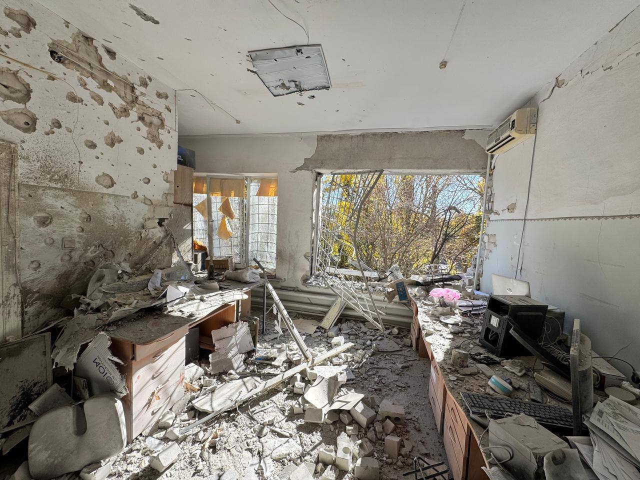 Зруйноване приміщення з численними слідами від осколків на стінах і великим проломом замість вікна. В кімнаті багато уламків, будівельного сміття, потрощених меблів та офісної техніки.