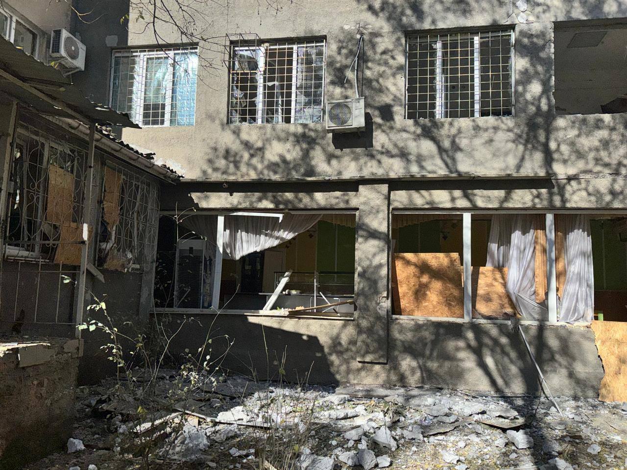 Фасад пошкодженої будівлі. Вікна першого поверху вибиті та частково закриті фанерою, а вікна другого поверху з металевими ґратами також пошкоджені. На землі видно уламки та сміття.