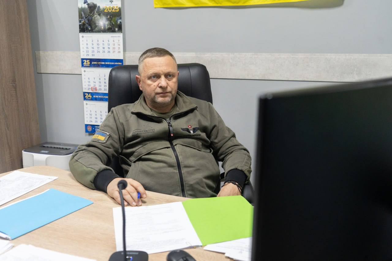 Чоловік у військовій формі оливкового кольору сидить за робочим столом з документами та монітором комп'ютера.